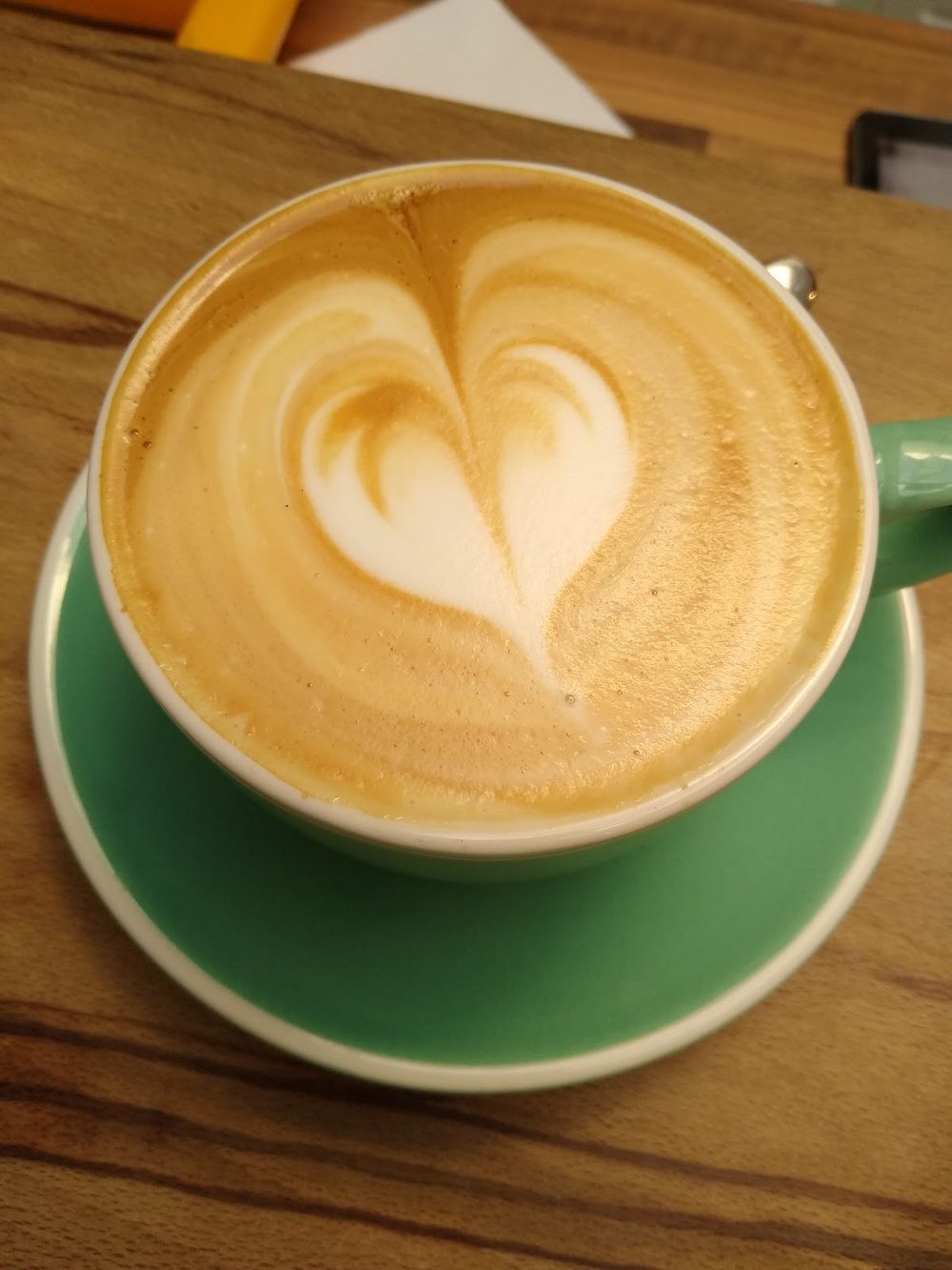 A latte with heart latte art in a green cup and saucer, shot from above on a wooden table